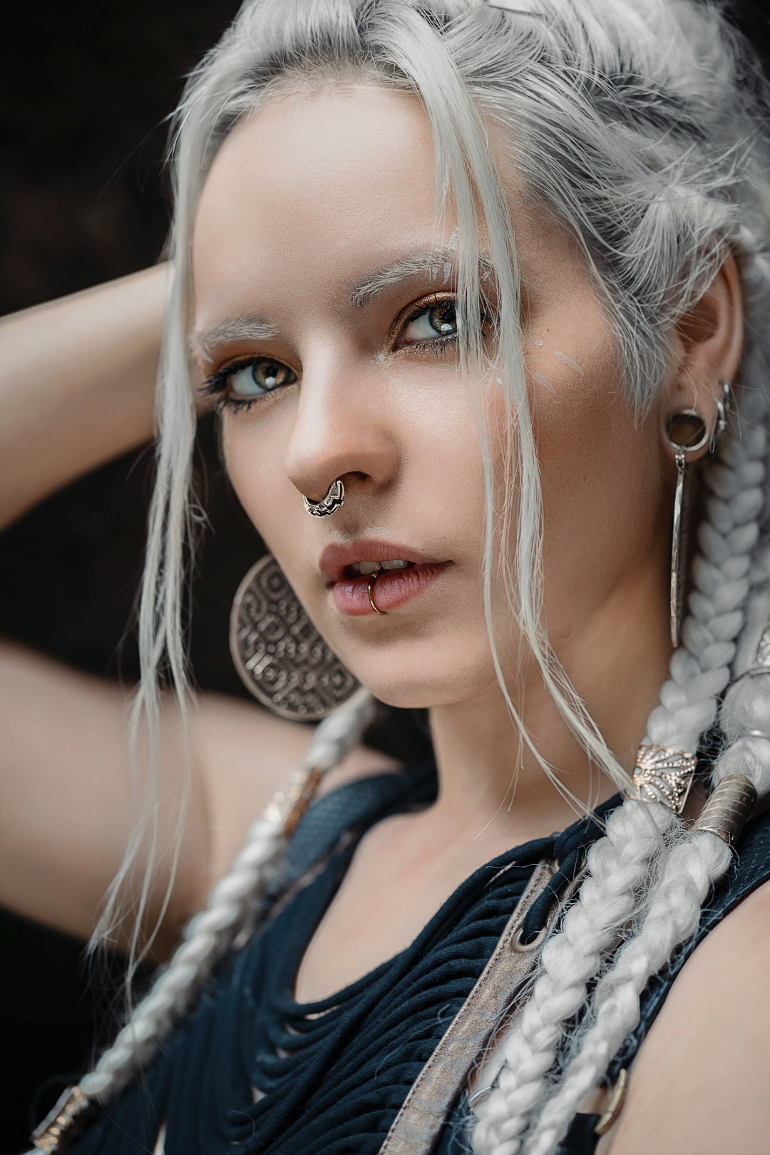 Close-up of a person with long white braided hair, subtle makeup, and striking light-colored eyes. They have an AYA Septum Clicker Shipibo Pattern Inspired by Silver 925 from Kaya Movement (16 gauge), multiple earrings, and are wearing a dark, textured top. Their expression is serious as they gaze directly into the camera.